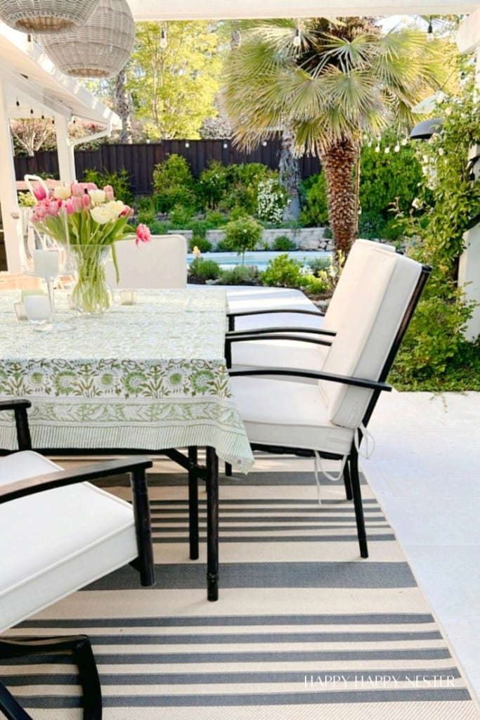 A cozy outdoor patio features a table covered with a floral-patterned cloth, topped with a bouquet of pink tulips in a vase. Four white cushioned chairs surround the table, all set on a striped rug. The summer decor is complemented by lush greenery, including a tall palm tree. Shop this look on Amazon!