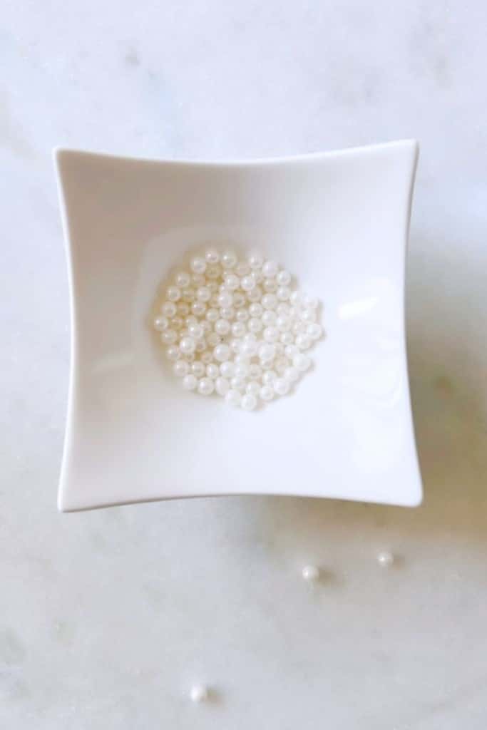 A small, square white dish holds several round pearls, neatly arranged. Two pearls rest outside the dish on the light-colored surface. The background has a soft, marbled texture, complementing the elegance of the pearls.