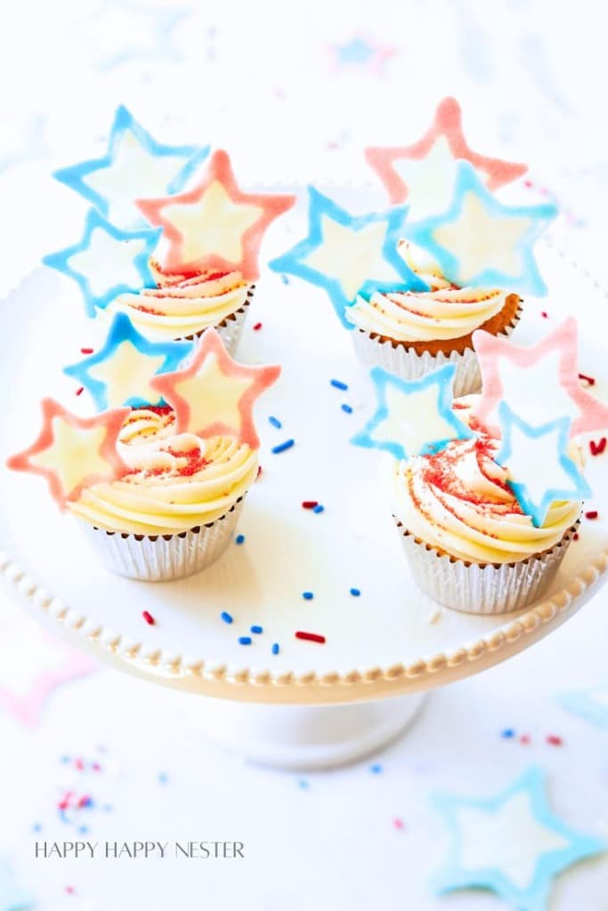 A white cake stand displays cupcakes topped with creamy swirls of white and red frosting. Each cupcake is adorned with star-shaped toppers in pastel blue and pink. Red, white, and blue sprinkles decorate the surface. Text reads "HAPPY HAPPY NESTER" in the bottom corner.