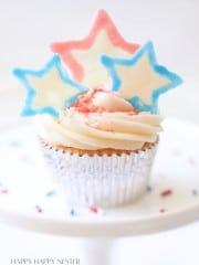 A cupcake with white frosting topped with three star-shaped decorations in blue, white, and red colors. The cupcake wrapper is a shiny silver, and it is sprinkled with red, white, and blue crumbs. The background is soft and out of focus.