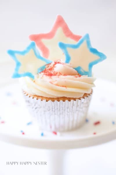 A cupcake with white frosting topped with three star-shaped decorations in blue, white, and red colors. The cupcake wrapper is a shiny silver, and it is sprinkled with red, white, and blue crumbs. The background is soft and out of focus.