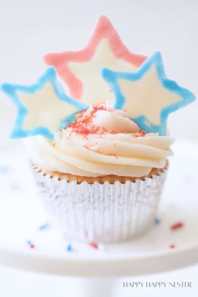 A vanilla cupcake in a silver wrapper, topped with white frosting and decorated with three star-shaped cookies in pastel colors (light blue, pink, and white). Red sprinkles are scattered on the frosting. The background is softly blurred.