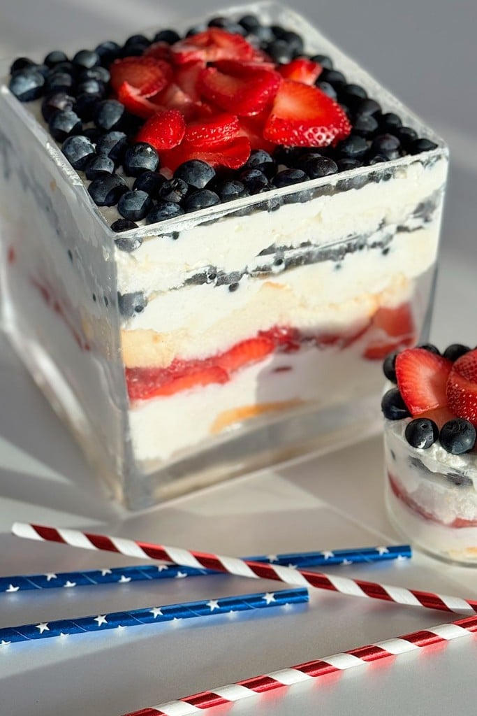 A square glass container filled with layers of whipped cream, mixed berries, and cake, topped with blueberries, sliced strawberries, and shards of white chocolate bark. A small portion is served in a glass cup alongside. Red, white, and blue striped straws are laid beside the dessert.