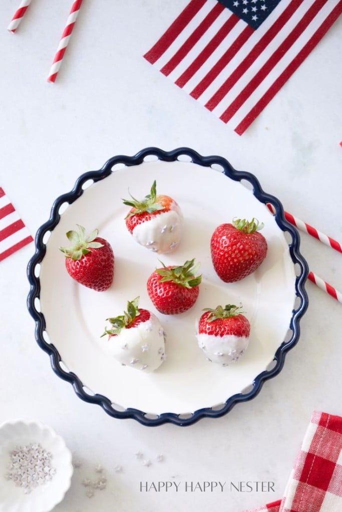 A decorative plate with a blue rim holds six fresh strawberries, three of which are partly dipped in white chocolate. The background features a white surface with red and white striped straws and a small American flag, giving a patriotic theme.