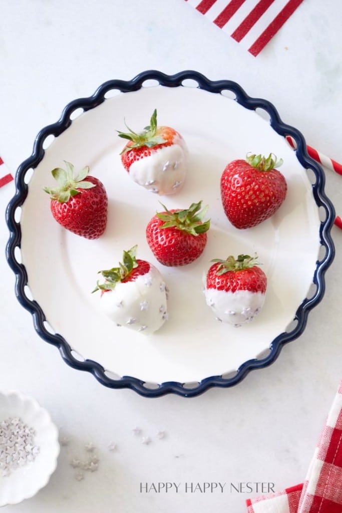 A white plate with a blue scalloped rim holds six strawberries, four of which are partially dipped in white chocolate. The plate is surrounded by red and white striped napkins and a partially visible bowl of salt on the left side.