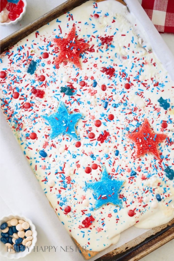 A festive sheet cake adorned with red, white, and blue sprinkles, star-shaped decorations, and coated with white frosting. Small bowls of red, white, and blue candies surround the cake, laid out on a white parchment-lined tray.