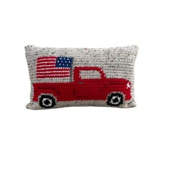A rectangular, knitted cushion with a design of a red pickup truck carrying a large American flag. The cushion's background is a light grey color with a textured knit pattern.
