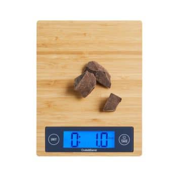 A digital kitchen scale with a bamboo surface, displaying a weight of 1.0 ounces. Several pieces of dark chocolate are placed on the scale. The display screen is illuminated in blue.