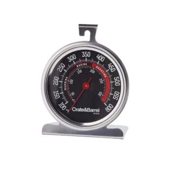 A round oven thermometer with a black face, red needle, and white markings showing temperature in Fahrenheit ranging from 100°F to 600°F. The brand name "Crate & Barrel" is displayed at the bottom of the dial. The thermometer has a hook and a stand.