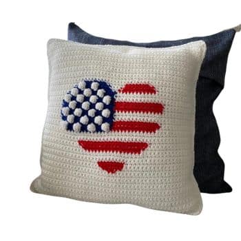 A square, white crocheted pillow featuring a heart design with the American flag pattern. The heart is composed of blue and white stars on the left and red and white stripes on the right. Another plain pillow in the background partially visible.