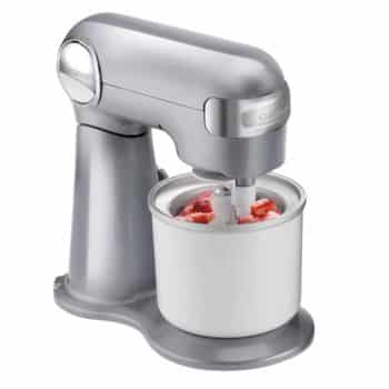 A stainless steel stand mixer with an ice cream maker attachment. The machine is churning ice cream, with pieces of strawberries visible inside the white ice cream bowl. The mixer has a sleek, modern design.