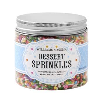 A clear plastic jar with a silver screw-top lid contains colorful dessert sprinkles. The label reads "Williams Sonoma Dessert Sprinkles" and mentions that it is used to decorate cookies, cupcakes, and other sweet treats. The net weight is 10 ounces.