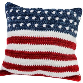 A square crocheted throw pillow featuring an American flag design. The top part is blue with white stars, and the bottom consists of red and white horizontal stripes.