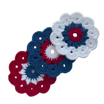 Four crocheted flowers are shown, each with rounded edges and a small hole in the center. The flowers are arranged diagonally. Their color schemes are red, white, and blue, featuring a mix of solid and patterned designs.