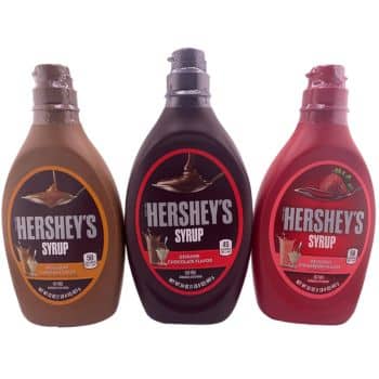 Three bottles of Hershey's Syrup, from left to right: caramel flavor in a tan bottle, genuine chocolate flavor in a dark brown bottle, and strawberry flavor in a red bottle. Each bottle features its respective syrup being poured onto a dessert or beverage.