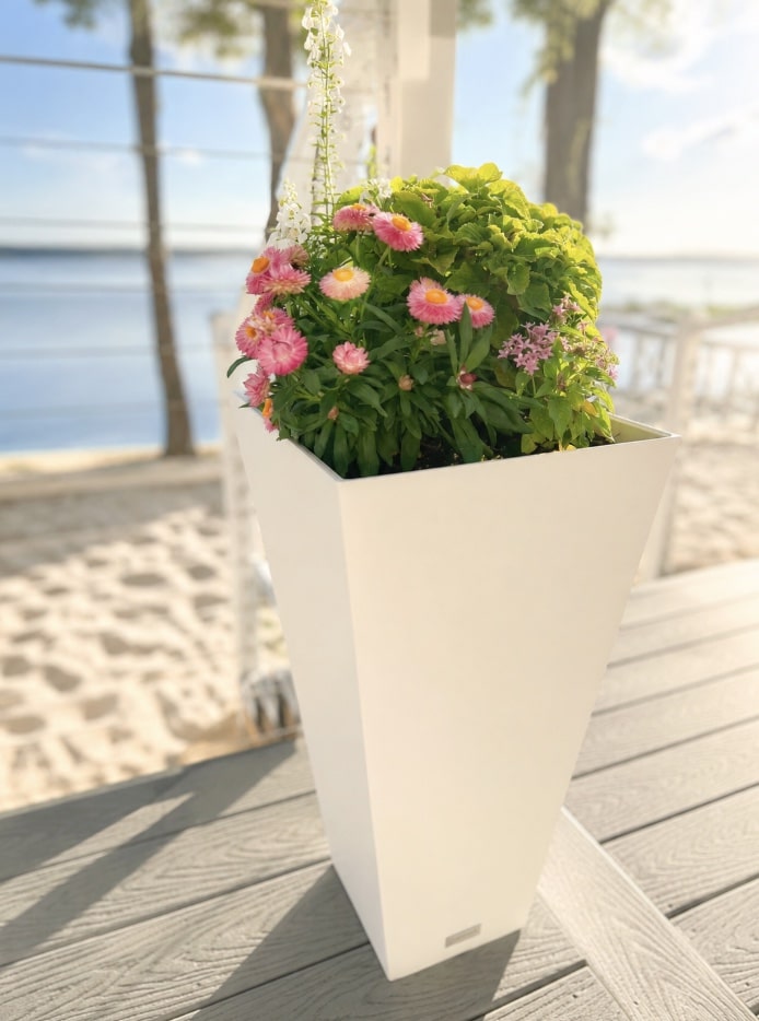 A tall, white planter with green foliage and pink flowers sits on a wooden deck near a sandy beach and calm water—perfect inspiration for flower garden decor ideas and more, surrounded by trees and sunshine in the background.