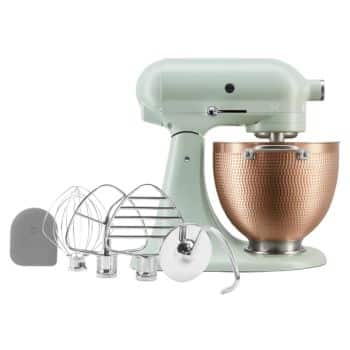 A mint green kitchen stand mixer with a copper mixing bowl is displayed against a white background. Various attachments, including a wire whisk, paddle beater, dough hook, and spatula, are placed in front of the mixer.