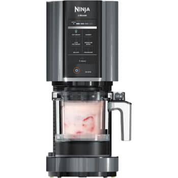 A black and gray Ninja CREAMi ice cream and sorbet maker, showing a partially visible touchscreen with various settings. The transparent container below contains a light pink frozen dessert mixture with chunks of strawberries inside.