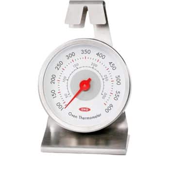 A stainless steel oven thermometer with a large circular dial displaying temperatures from 100 to 600 degrees Fahrenheit. The thermometer has a red needle and the brand name "OXO" in red at the center. It features a built-in clip for easy attachment inside the oven.