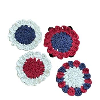 A set of four crocheted coasters arranged in a cluster. Each coaster features a circular design with alternating colors of red, white, and blue yarn. The intricate patterns and scalloped edges give the coasters a handmade, decorative appearance.