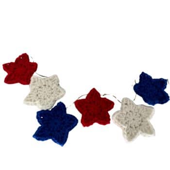 A string of crochet stars in red, white, and blue colors are arranged in a line. The stars are alternating in color and connected with twine. The design is festive and patriotic, often associated with celebrations or decorations for occasions like Independence Day.
