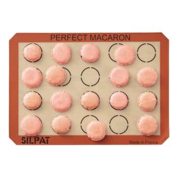 A tan-colored baking mat with a red border labeled "SILPAT" and "PERFECT MACARON." The mat has 15 baked pink macarons evenly spaced within circles printed on the surface. The mat is labeled "Made in France" at the bottom corner.