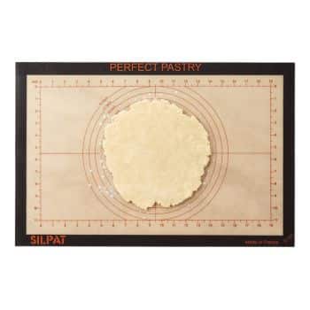 A round piece of dough sits on a Silpat "Perfect Pastry" baking mat. The mat has measurement guides and concentric circles for precise pastry shaping. The mat's edges are black with "PERFECT PASTRY" written in red at the top and "SILPAT" at the bottom left.