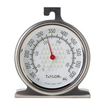A Taylor oven thermometer displaying temperature readings in both Fahrenheit (0 to 600) and Celsius (50 to 300). The needle is pointing to approximately 250 degrees Fahrenheit. The thermometer features a stainless steel frame and a stand for stable placement.