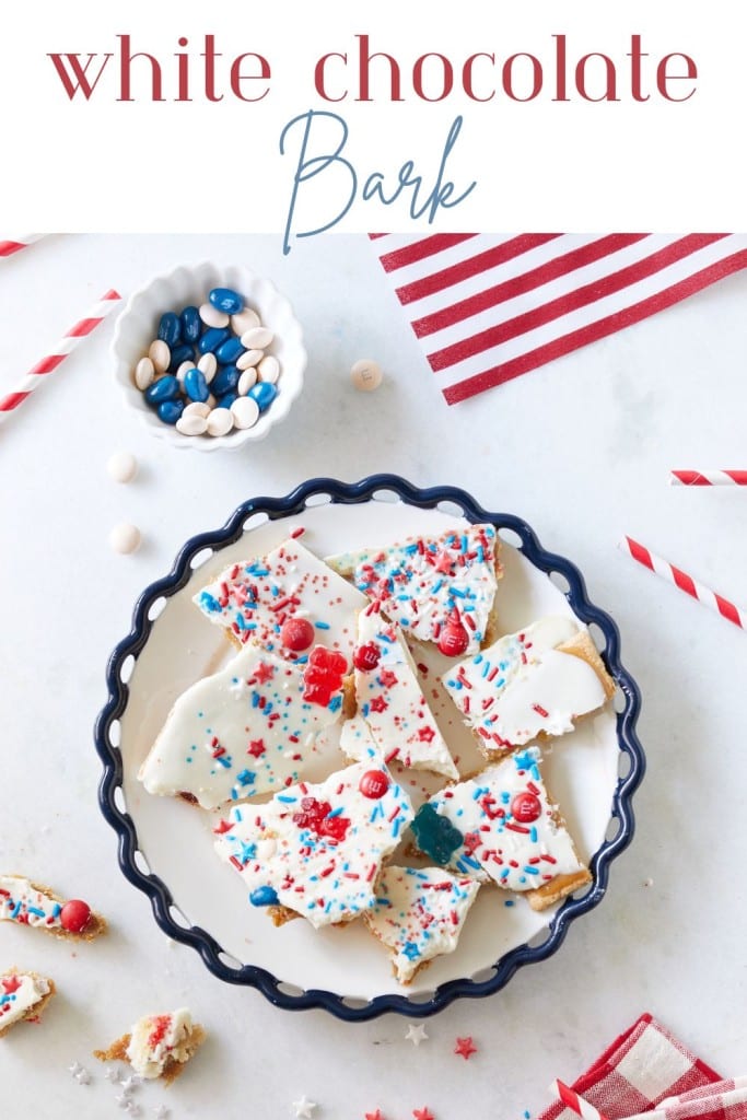 A decorative blue-edged plate holds pieces of white chocolate bark topped with colorful sprinkles and red, white, and blue candies. In the background, there are red and white straws, a small bowl of additional candies, and festive decorations. "White Chocolate Bark" is written in the top area of the image.