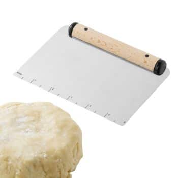 A metal dough scraper with a wooden handle lies on a white surface. The scraper has measurement markings along its edge. In front of the scraper, there is a ball of dough dusted with flour.