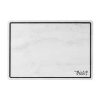 Image of a rectangular marble cutting board with a black border. The bottom right corner is branded with the text "Williams Sonoma." The marble pattern is primarily white with light gray veining.