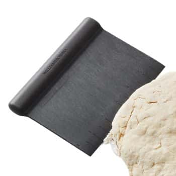 A black dough scraper with measurement markings is shown partially cutting through a ball of dough. The handle of the scraper has the brand name "Tupperware" engraved on it. The background is plain white.
