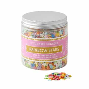 A clear jar with a silver lid containing star-shaped sprinkles in various rainbow colors. The jar has a pink label that reads "Williams Sonoma Rainbow Stars" with additional small text below it. A small pile of the sprinkles is shown outside the jar.