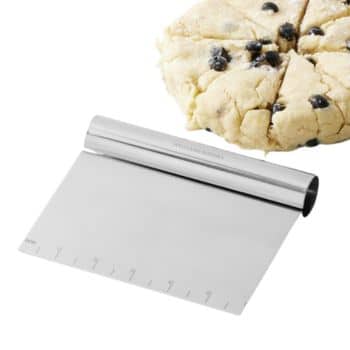A dough scraper with a silver handle and measurement markings along the edge is placed in front of a partially sliced, round blueberry scone dough.