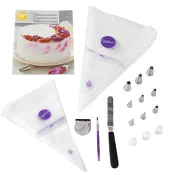 A cake decorating kit including a booklet with a cake on the cover, two icing bags, various decorating tips, a purple brush, a cake comb, a spatula, and purple and white accessories. The items are spread out on a white background.