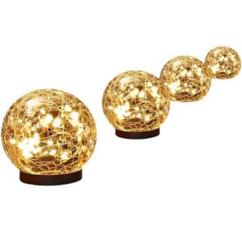 Four glowing spherical table lamps of descending size are arranged in a line. Each lamp has a cracked glass design that sparkles with warm, yellowish light. Perfect for flower garden decor ideas, the lamps are placed on black circular bases.