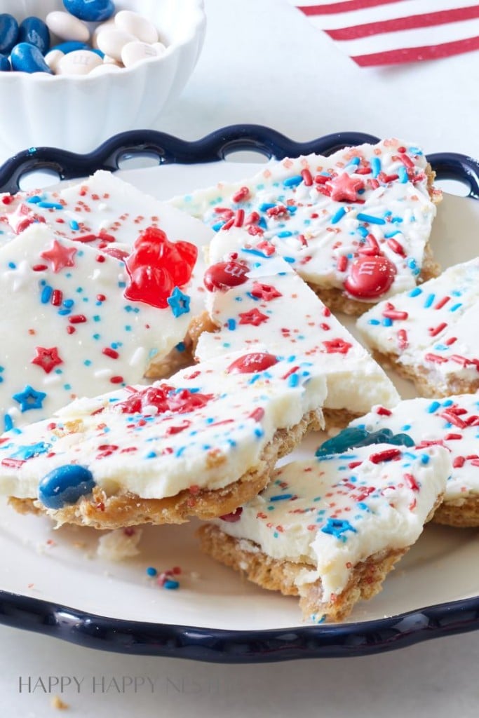 A plate with several pieces of festive white chocolate bark adorned with red, white, and blue sprinkles and star-shaped candies. In the background, a bowl filled with red, white, and blue M&M's is partially visible. The bark has a light, crispy texture.
