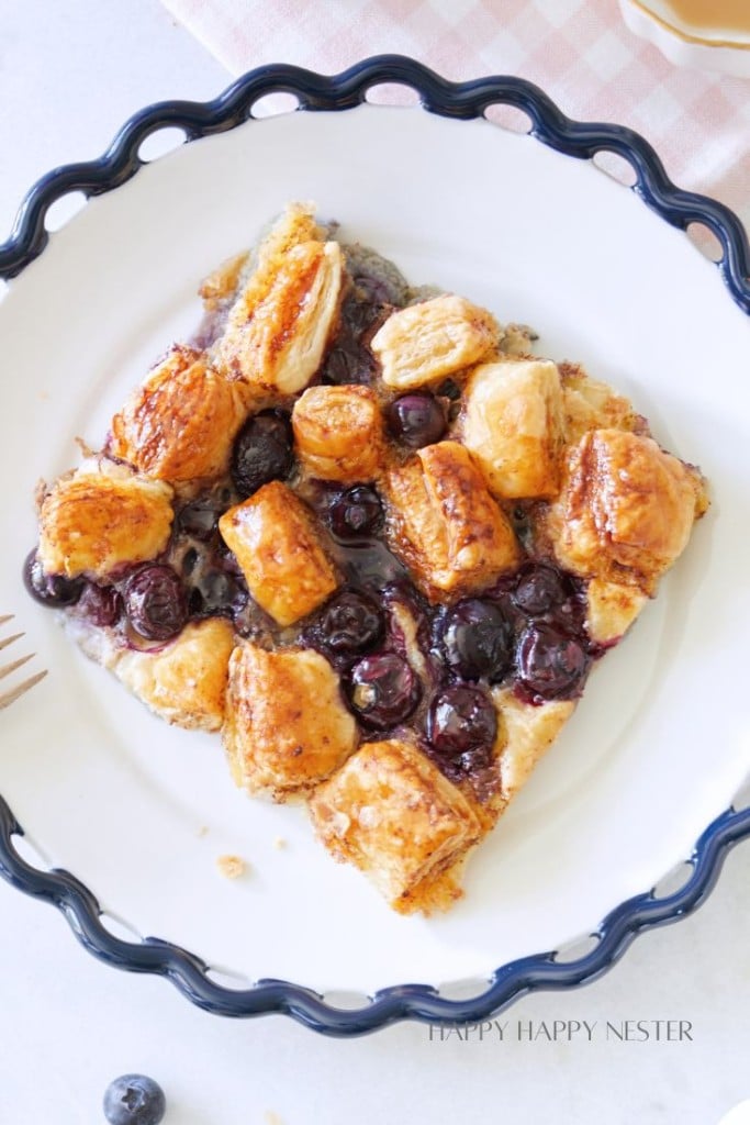 A white plate with a decorative rim holds a slice of blueberry puff pastry tart. The tart features golden-brown flaky pastry squares and juicy blueberries, all glazed with a shiny coating. Part of a fork and a lone blueberry are visible near the plate.