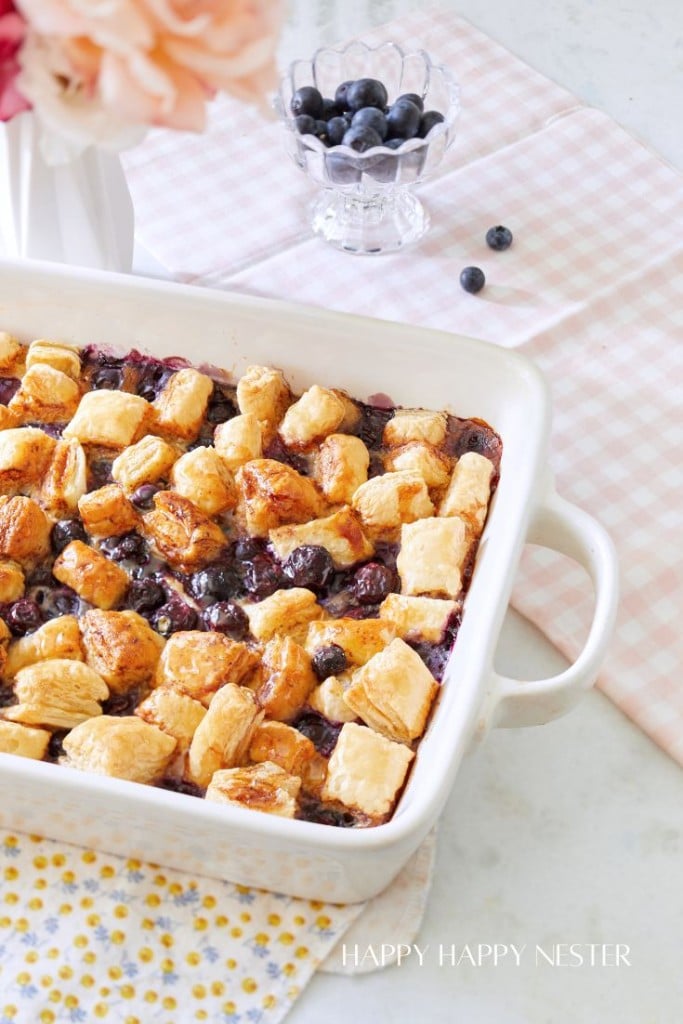 A baking dish filled with freshly baked blueberry cobbler, topped with golden, flaky pastry. The dish is placed on a light checkered cloth with a glass bowl of blueberries nearby. Pink flowers in a vase are partially visible in the corner.