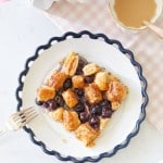 A plate with a wavy edge holds a pastry topped with blueberries and glazed puff pastry squares. A fork rests beside the plate. Two eggs, some loose blueberries, and a peach-colored, scalloped dish with coffee sit nearby on a pink checkered tablecloth. Flowers are in the top corner.