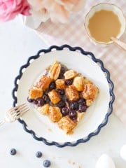 A plate with a wavy edge holds a pastry topped with blueberries and glazed puff pastry squares. A fork rests beside the plate. Two eggs, some loose blueberries, and a peach-colored, scalloped dish with coffee sit nearby on a pink checkered tablecloth. Flowers are in the top corner.