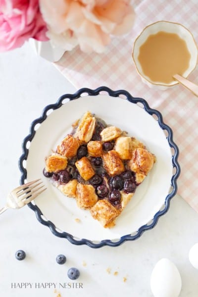 A plate with a wavy edge holds a pastry topped with blueberries and glazed puff pastry squares. A fork rests beside the plate. Two eggs, some loose blueberries, and a peach-colored, scalloped dish with coffee sit nearby on a pink checkered tablecloth. Flowers are in the top corner.