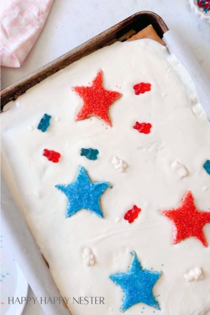 A rectangular cake on a baking sheet is decorated with white frosting, red and blue star-shaped sprinkles, and small red, white, and blue gummy bears. The baking sheet is lined with parchment paper, and a pink and white cloth is partially visible in the upper left corner.