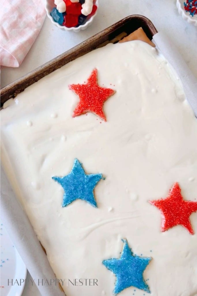 A frosted sheet cake topped with three star-shaped decorations: two red and one blue, all covered in colored sugar. The cake sits in a baking tray lined with parchment paper. In the background, a small bowl with red and white candies is partially visible.