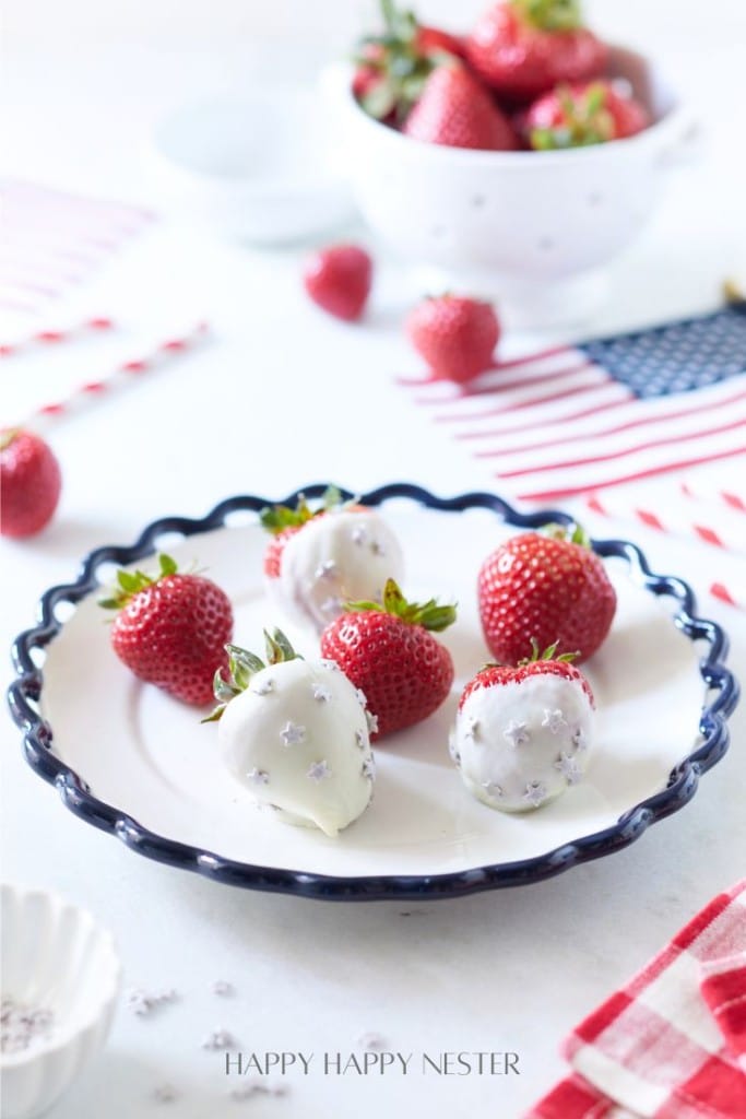 A white plate with a blue-rimmed edge holds five strawberries, two of which are coated in white chocolate with sprinkles. In the background are a white bowl filled with more strawberries, an American flag, and red-and-white striped straws on a light-colored table.