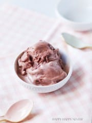 A small white bowl filled with scoops of chocolate ice cream is placed on a light pink and white checkered fabric. Two spoons, one gold and one pink, rest beside the bowl. The background is softly blurred with a ceramic bowl visible.