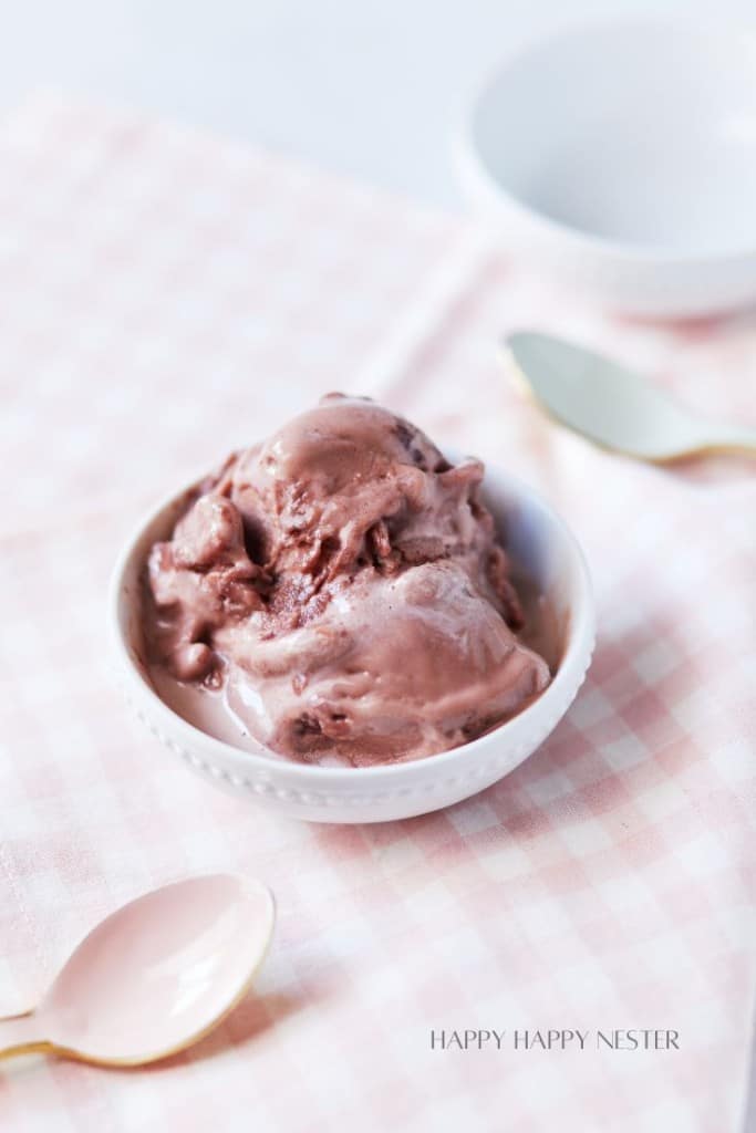 A small white bowl filled with scoops of chocolate ice cream is placed on a light pink and white checkered fabric. Two spoons, one gold and one pink, rest beside the bowl. The background is softly blurred with a ceramic bowl visible.