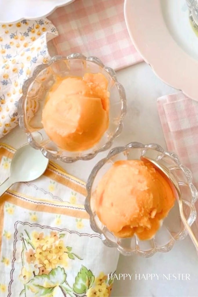 Two scoops of orange sherbet are served in clear glass bowls. The bowls are set on a table with floral and checkered napkins, one of which contains a spoon. The scene is bright and cheerful, with pastel colors dominating the accessories in the background.