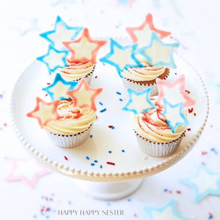 Four cupcakes topped with red, white, and blue star-shaped decorations are displayed on a white, beaded-edge cake stand. The cupcakes have creamy white frosting with colorful sprinkles scattered around. The background is softly blurred with more star-shaped decorations.