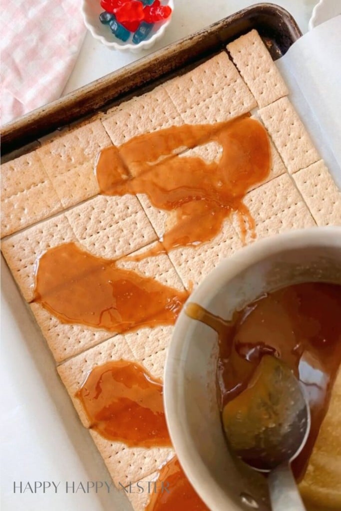 Image shows a baking sheet lined with graham crackers, partially covered with caramel sauce being poured from a bowl. In the background, there is a small dish with colorful gummy candies. A pink and white checked cloth is partially visible on the side.